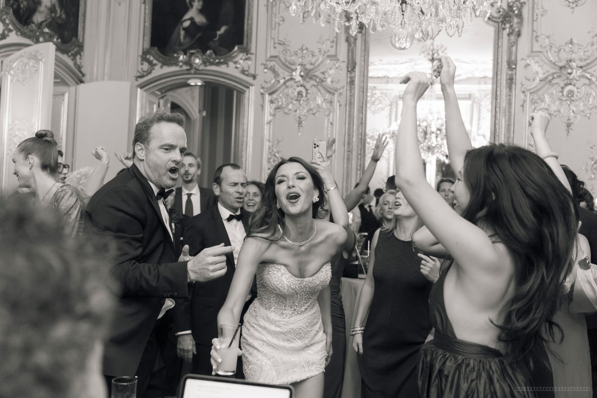 Les mariés et leurs invités célèbrent dans une salle aux miroirs et lustres, en noir et blanc
