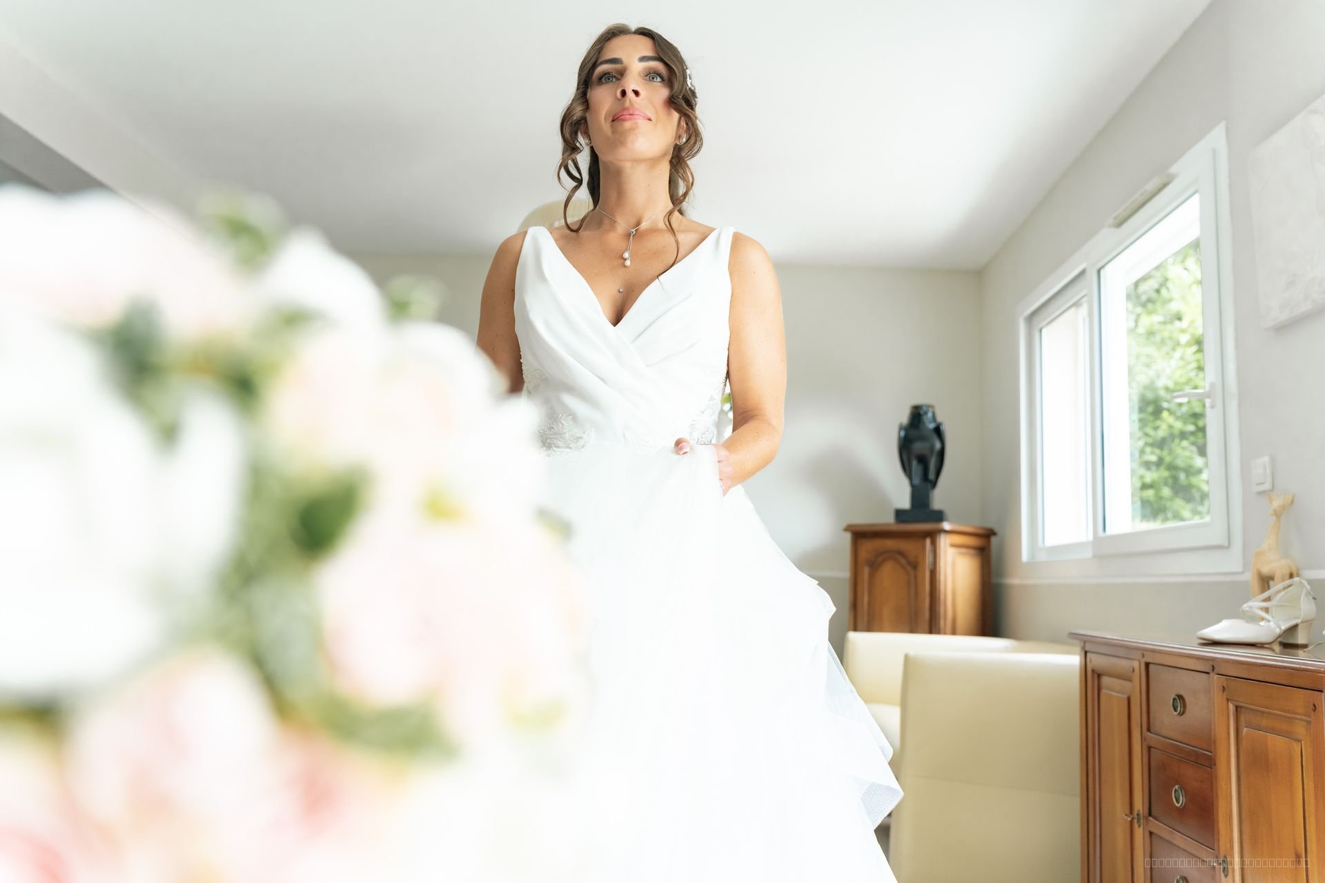 La mariée debout dans une chambre lumineuse, un bouquet flou au premier plan