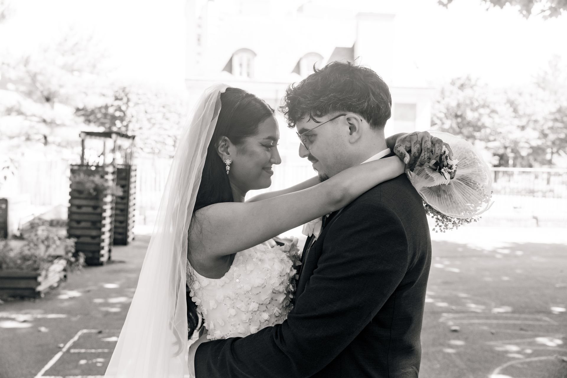 Les mariés s’enlacent en se regardant, la mariée tenant son bouquet derrière le marié, en noir et blanc