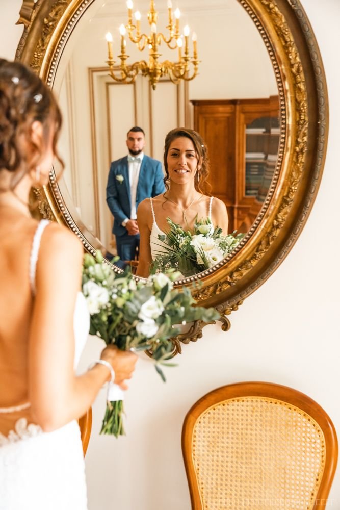 Reflet de la mariée tenant son bouquet dans un miroir doré, le marié en arrière-plan