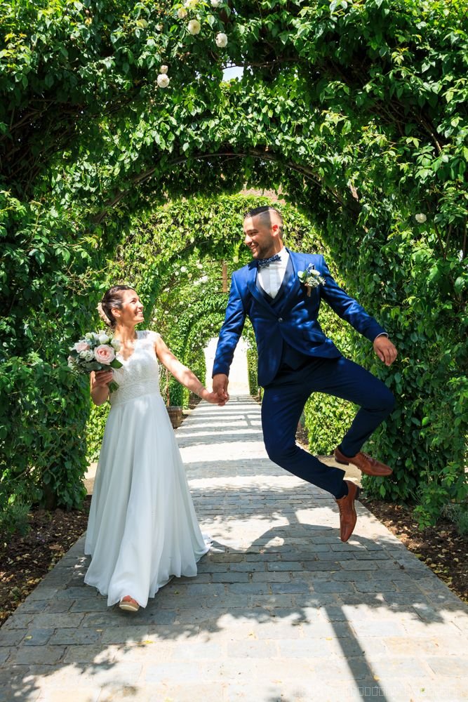 Le marié saute en tenant la main de la mariée dans une allée bordée d’arbres
