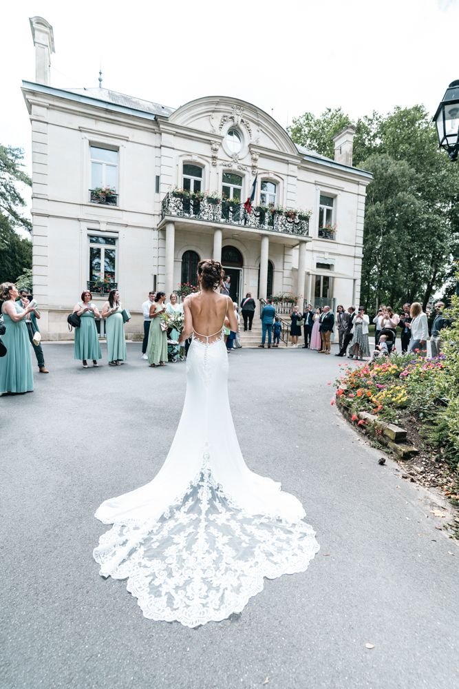 La mariée de dos, sa traîne en dentelle déployée devant les invités réunis à l’extérieur