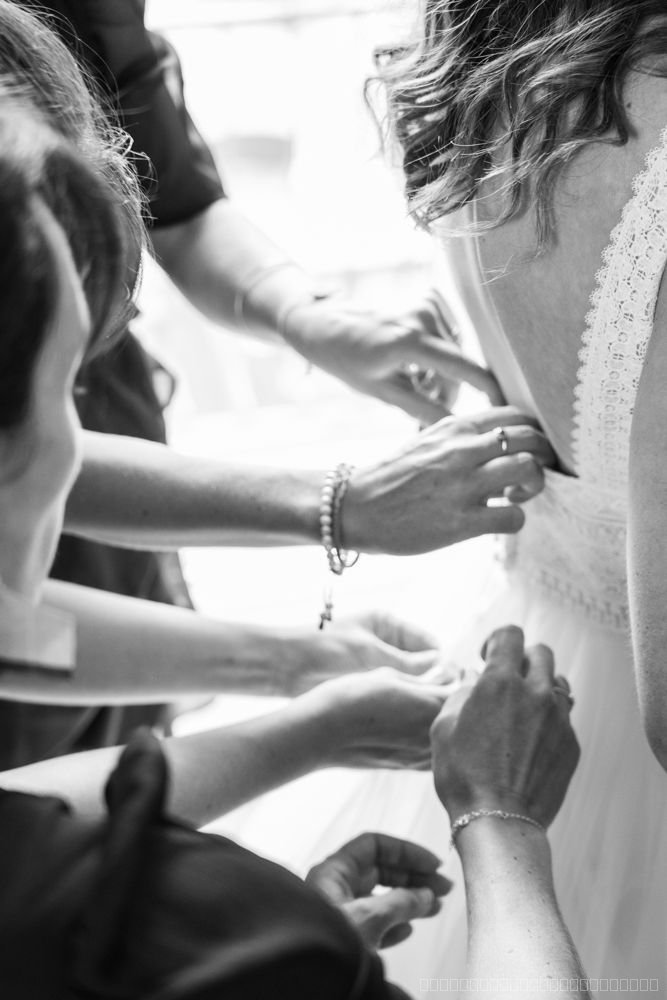 Plusieurs mains ferment le dos de la robe de mariée pendant les préparatifs, en noir et blanc
