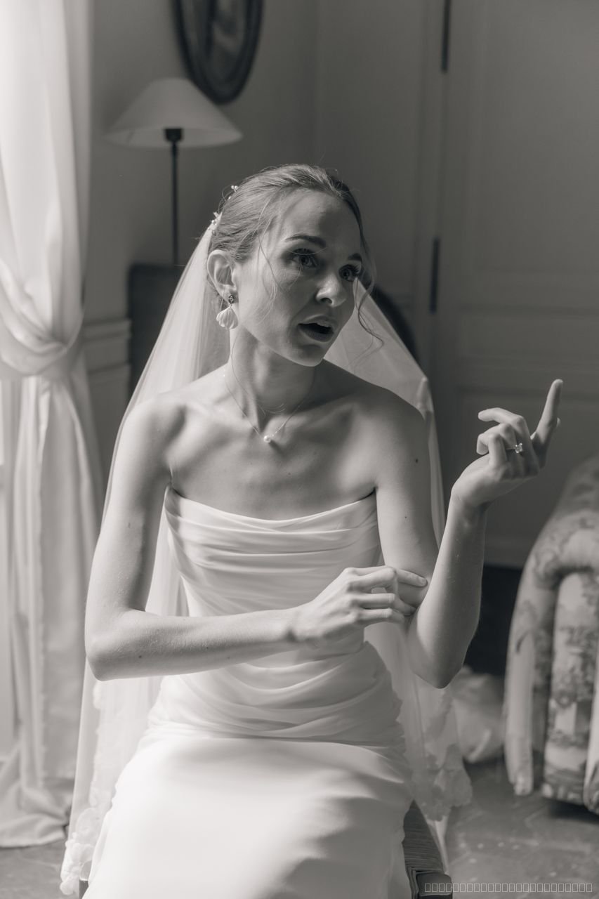 La mariée assise près d’une fenêtre fait un geste de la main, photographiée en noir et blanc