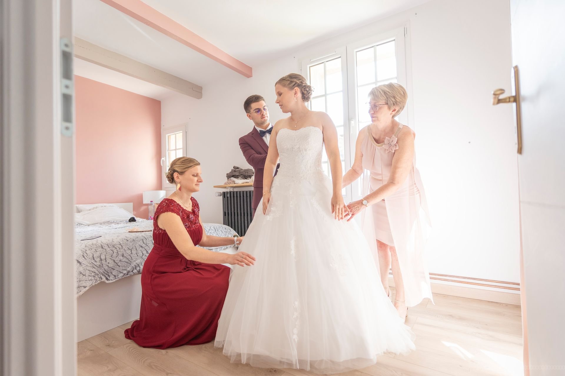 La mariée entourée de plusieurs personnes qui ajustent sa robe dans une chambre lumineuse